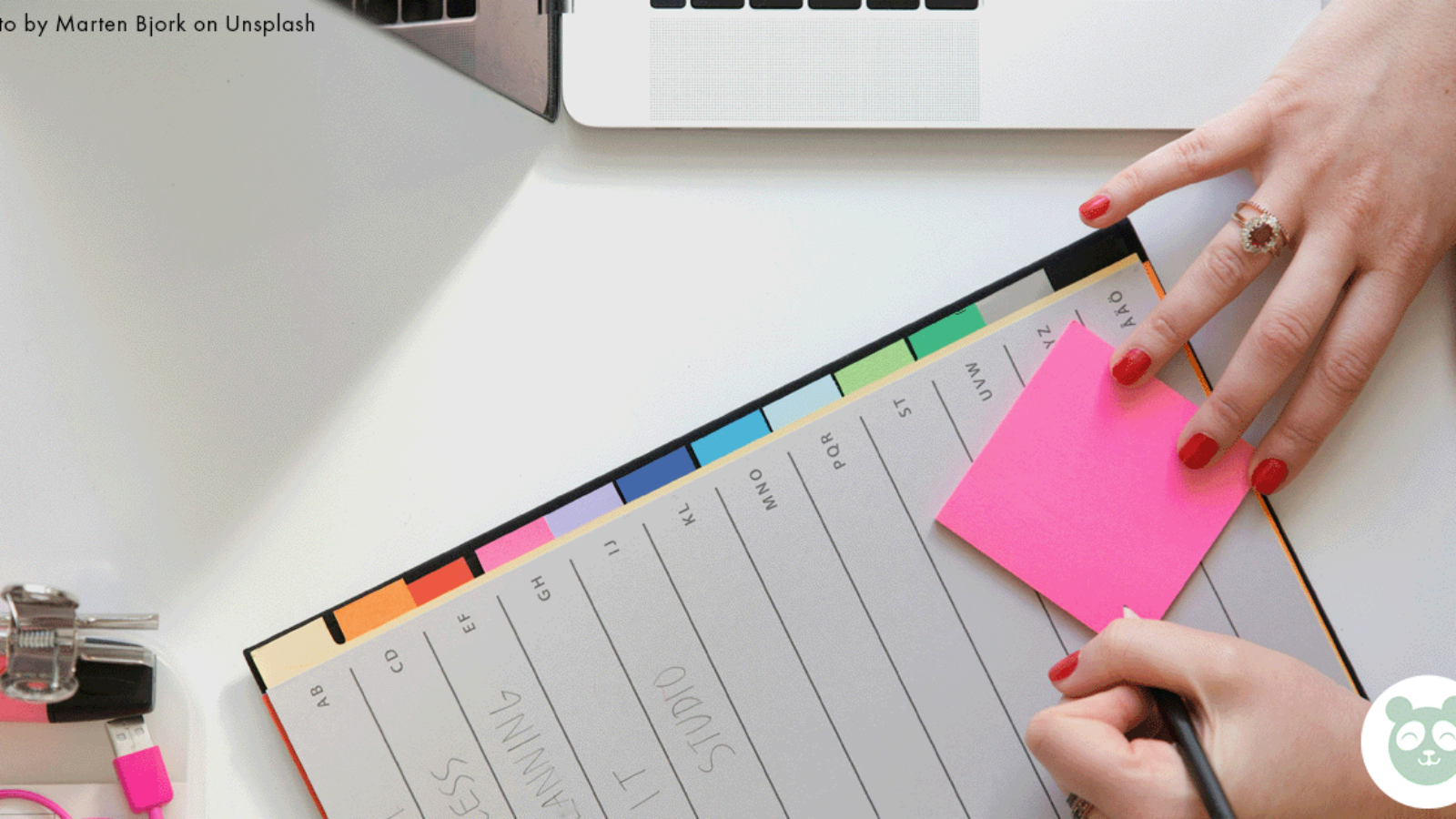 A large planner/diary. A Ladies hands with red nails writing on a pink post it note